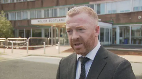 DCI Wright is wearing a grey suit and tie. He has short ginger hair and a cropped ginger beard. He is standing in front of Police Scotland headquarters at Fettes in Edinburgh.