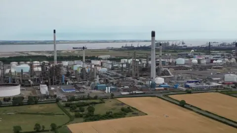 A drone view of the Prax Lindsey Oil Refinery. A cluster of metal structures rising to the grey sky. Green and yellow stubble fields can be seen. 
