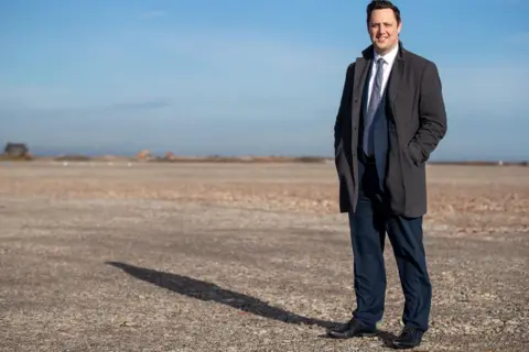 Tees Valley Combined Authority Ben Houchen, dressed in a navy suit and grey coat, looks into the camera. He has short black hair.