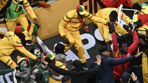 A scene of crowd disturbance at the 2025 Afcon final in Rabat sees multiple Senegal supporters, all dressed in yellow, green and red, coming over a barrier and clashing with security. One man is wielding a white chair above his head while two of the security staff members have their arms outstretched with palms up in a bid to calm proceedings