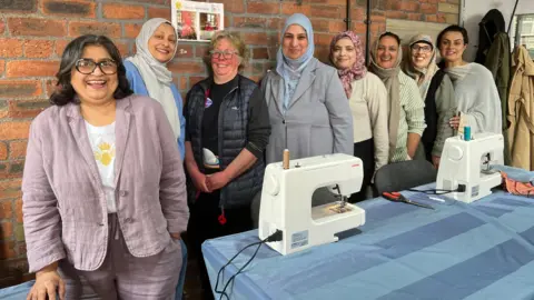 Aisha Iqbal/BBC A row of ladies, of different backgrounds and ages, pose behind a table full of sewing machines and equipment.