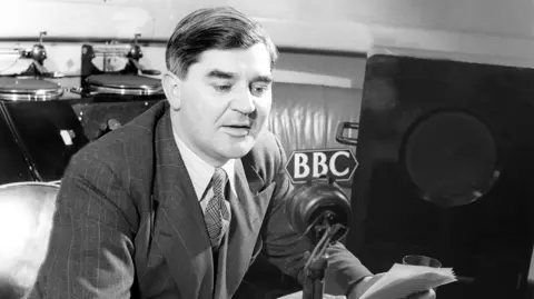 A black and white image of Nye Bevan delivering a broadcast on the radio. The microphone is branded with the BBC logo.