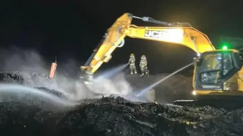 A yellow crane can be seen to the right, its arm out over a sea of scorched and blackened waste, while the beams of its light in the dark show two firefighters in yellow high-viz using a hose to help dampen the fire. Smoke is seen floating upwards from the scene. 