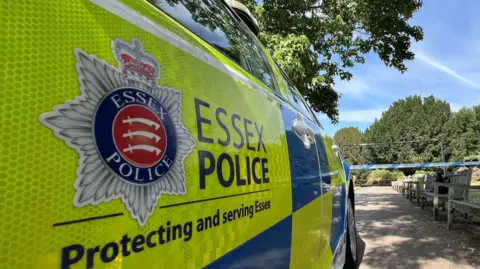 An Essex Police car parked in front of police tape. There are trees and bushes in front of the car.