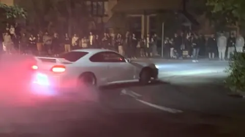 A white car drifting on a road with people surrounding it