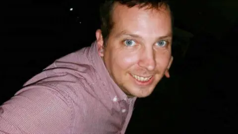 Family handout A man is leant forward into the middle of the frame, and is looking at the camera. He is smiling and wearing a red shirt. The background is black.