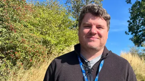 A well-built man with short, thick brown hair and stubble looks towards the camera as he stands in countryside, surrounded by long grass and green shrubs under a blue sky. He is wearing a black fleece, grey T-shirt and a blue "staff" lanyard.