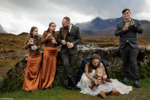 لین کندی.  خانواده ای در حالی که کنار دیواری در جزیره اسکای اسکاتلند ایستاده اند کیک می خورند