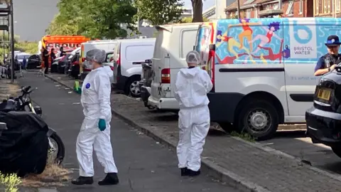 Two people in white outfits walk down a residential street past parked cars