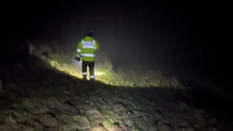 Swanage Coastguard A man from HM Coastguard wearing hi-vis clothing is pictured from behind, searching through the darkness with a torch. 