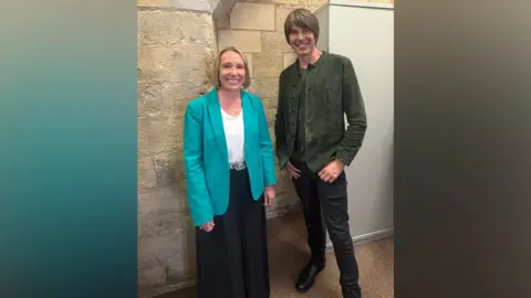 Helen Morgan MP On the left, there is a woman in a turquoise-teal blazer, white t-shirt and black trousers. She has blonde ear-length hair and is smiling. On her right, there is a man with black jeans, black trousers and a dark grey jacket. He also has brown hair brushed to his left.