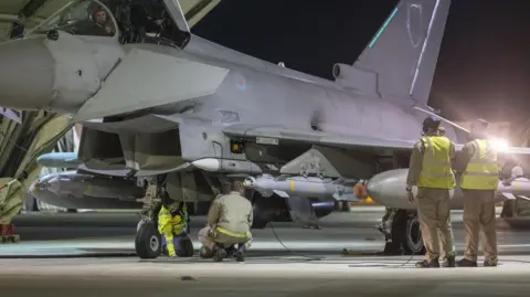 Royal Air Force ground crew preparing an RAF Typhoon fighter jet. It is night-time and the team are working under the floodlights of a hangar.
