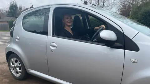 Young woman wearing a black long sleeve top sitting in a silver car and smiling at the camera. 