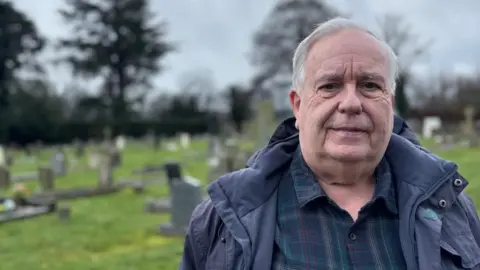 BBC Man in waterproof jacket and a navy shirt stood in a graveyard with several stone headstones. He has grey hair and a serious expression. 