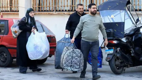 A woman and two men walk along a street in Beirut carrying big bags of belongings (03/03/26)