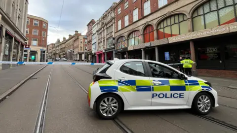 A cordon in Cheapside, Nottingham city centre. Police tape blocks tram lines and a police car is parked across the road