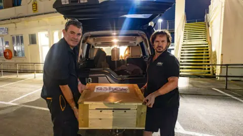 Chris George Two men holding a GSPCA wooden box either side. They are putting it into the boot of a vehicle. 