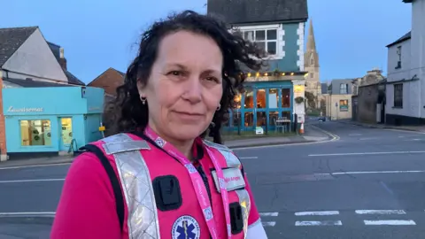 A woman with dark curly hair wearing a pink long sleeved top and pink high-vis jacket is smiling at the camera. She is wearing a badge which says 'Night Angels' and also a bright pink lanyard.