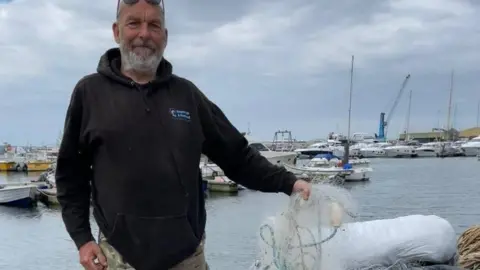 Weymouth & Portland Marine Litter Project Volunteer Dave Taylor on Poole harbourside with a handful of white gill netting with various boats on the water behind him