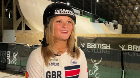 A woman with long blonde hair, a black helmet and white British Cycling top, sits and smiles. Behind her is a British Cycling barrier with a ramp behind it.