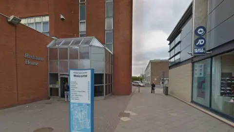 A brick-built block of flats with several storeys, with a glass entrance porch and "Redstone House" in silver lettering on a brick wall at right angles to the main building. There is a blue and white information map on a sign in the foreground. There is a two-storey JD Sports store to the right. There is a pavement between the two building on which a person is standing.