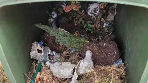 En un contenedor de basura general, junto con montones de césped, yacían adornos de Louise Hirst, entre los que se incluían un par de palomas y flores artificiales.
