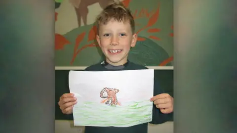 Twyford Primary School A boy is pictured smiling at the camera while holding a hand drawn picture of a capybara in both hands. 