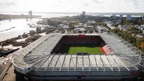 An aerial view of St Mary's Stadium, which is rectangular with a large white roof over seating on all four sides. Beside the football ground, bright sunlight reflects off the River Itchen which opens into the Southampton Water estuary in the background. Machinery and piles of aggregate materials can be seen beside the river.