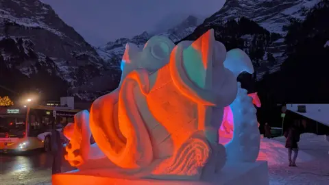 Red and pink lights on a snow sculpture of a sea monster and a ship with snow covered mountains and a bus in the background