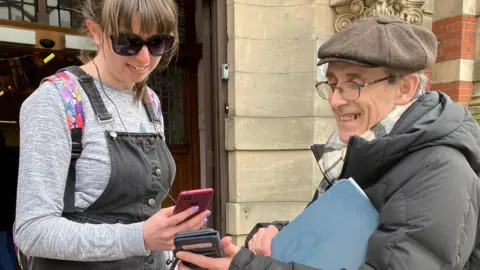 A man on the right in a black puffer coat, grey and white striped scarf and brown tween flat cap smiling as he shows his phone to a woman, who is scanning it with her own. The man has wire-rimmed glasses and is standing in the street outside a sandstone building. The woman has dark hair tied back and is wearing sunglasses, a grey long-sleeved top and black denim pinafore.