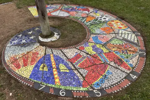 Large multi-coloured mosaic on the ground with images of boots, a football, stars and other items. It has numbers on the side and is spiral shaped. It has a post in the middle sticking out of the ground. 