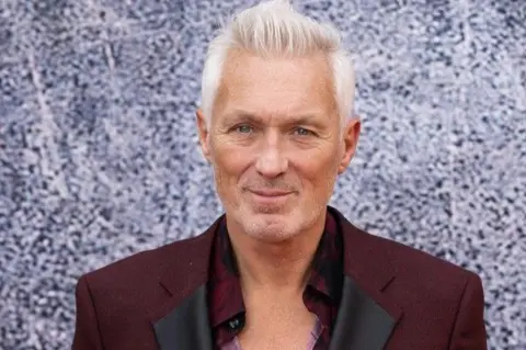 Getty Images Martin Kemp, who has short grey hair combed up, stares directly into the camera at a movie premiere. He is wearing a maroon open-necked shirt and a maroon blazer. 