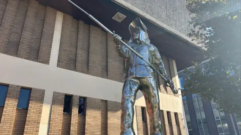 A metal statue showing a life-sized man dressed in steelworkers' uniform and holding a long pole with both hands.