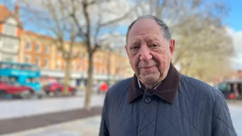 Jeremy Nettle wearing a navy quilted jacket with a brown collar. He is smiling at the camera. The background is blurred but shows a street with cars, shops and a line of trees.