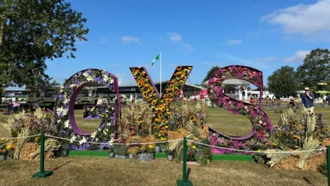 BBC/CATHY KILLICK A picture of big metal sculptures of the letters 'G,' 'Y' and 'S.' Flowers are embedded within the letters and more flower displays, on top of hay bales, and on each side of them. A band-stand and a ferris wheel are in the background.