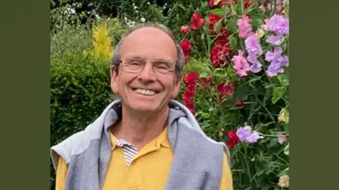 Stephen Stephen, who has short grey hair, back from a receding hairline, is wearing glasses and is smiling broadly. He has a grey sweatshirt over a yellow top and is standing in front of a hedge with red, pink and mauve sweet peas growing on his left.