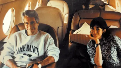 US Department of Justice Jeffrey Epstein and Ghislaine Maxwell sitting on either side of the aisle of what appears to be a small private plane, with cream leather seating and porthole windows. Epstein wears a grey sweatshirt which says "The Ranch" across it as he smiles broadly at the camera, while Maxwell wears a blue and white shirt and rests her chin on her palm as she smiles.