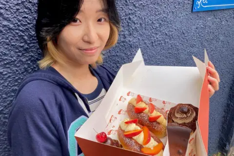 Wendy has black and dyed yellow hair. She is smiling at the camera and is wearing a blue hoodie. She has a big box of cakes in her arms.