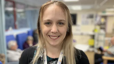 Danielle Bagg, deputy divisional medical director for medicine, Salisbury District Hospital is standing smiling in front of patients in a waiting area on the Acute Medical Ward.