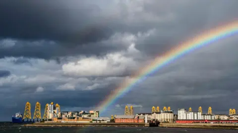 David Henderson A rainbow over a seaside town, there is a ship to one side, and water in the foreground, under a dark sky.