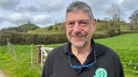 Man with grey hair who is smiling at the camera. Glastonbury Tor is the background with fields in the foreground. He has blue glasses hanging around his neck and a black t-shirt on with a green logo saying "Festival Medical Services".