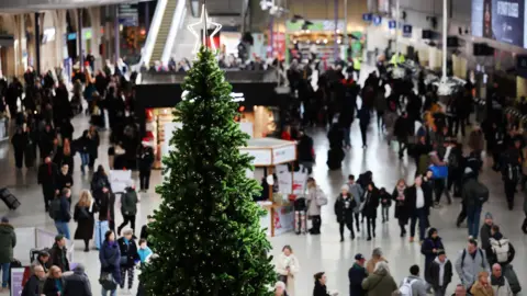 Commuters make their way home on 'Mad Friday', the last Friday before Christmas Eve, at Waterloo Station. There is a christmas tree with a star topper in the centre-left of the image and lots of commuters.