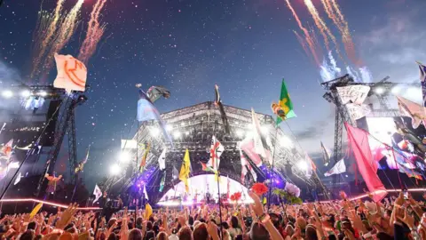 Crowds gathered around Glastonbury's Pyramid stage at night time. There are fireworks shooting up into the air and some people are waving flags around.