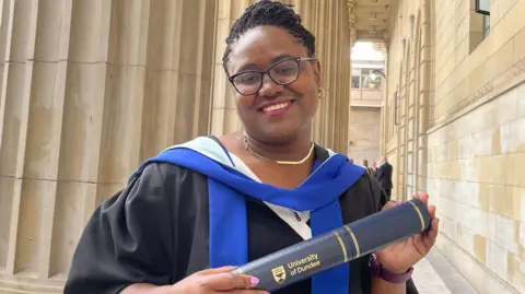 Police Scotland Dr Gomo, who is wearing black graduation robes, smiles as she holds a University of Dundee scroll. She is also wearing glasses.