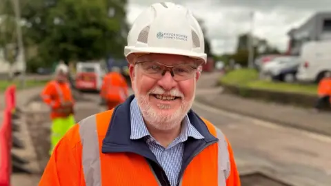 Richard Lovewell has a short white beard and is wearing goggles and a white hard hat. He also has an orange hi-vis on over a blue and white checked shirt. Behind him, contractors are fixing potholes.