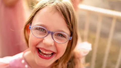 Leanne Rinne/BBC A young girl with blonde hair wearing pale purple rimmed glasses and smiling into the camera.