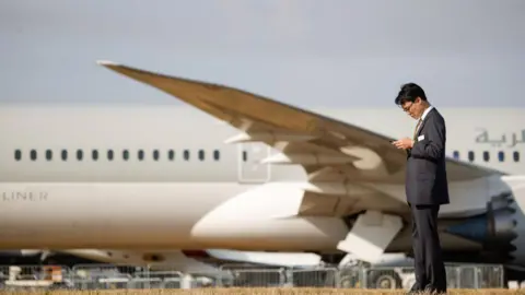 A man in a black suit looks at his phone while behind him is parked a Boeing 787-10 Dreamliner aircraft, operated by Qatar Airways QCSC, on the opening day of the Farnborough International Airshow in Farnborough, UK, on Monday, July 18, 2022. 