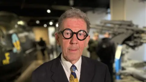 William Tate, an older gentleman with short, greying hair, wearing round large black-framed glasses. He is also wearing a dark suit, a white shirt, and a patterned tie featuring gold and blue designs. 