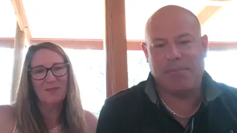 A woman with long dark-blonde hair and glasses and a bald man wearing a black collared shirt and a chain necklace sit next to each other in a screengrab from a video call.