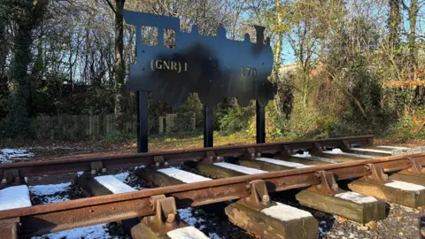 A sculptural silhouette of a train sits alongside a railway track. The depiction of the train bears the mark (GNR) 1 170 in gold lettering. The wooden rail sleepers have a sprinkling of snow upon them. Behind is a fenced garden with trees circling the background. 
